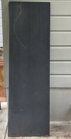 Full view of the large black slate slab leaning against a wall, showing its rectangular shape, length, and surface texture.