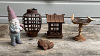 Overview of all five items together on wooden surface: garden gnome, two lanterns, bird fountain with two birds, and cast iron frog figurine.