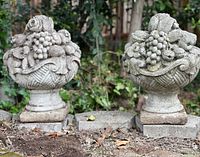 Front view of pair of antique cast stone garden urns with detailed fruit basket designs, showing weathered patina and slight damage to one urn.