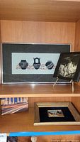 Framed art textile with three vases and a black-and-white signed photo art on wooden shelf