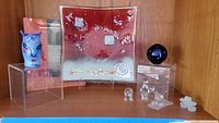 Full view of shelf with all glass items: square decorative plate, blue round glass vase, three small clear glass figurines (two penguins, one person), and frosted abstract figure.