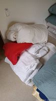 White comforter, red blanket and white linens stacked on the floor near a bed.