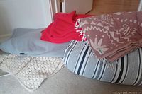 Five blankets stacked on the floor showing variety of colors and patterns: gray fleece, red fringe, pink geometric, blue and white stripes, and chunky knit cream