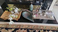 Photo showing the bamboo tray, elongated leaf-shaped bowl, faux lilies, and four metal candle holders arranged on a black surface in front of a fireplace.