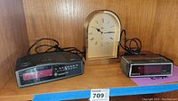 Photo showing all three clocks: Howard Miller mantle clock, Spartus alarm clock, and General Electric clock radio on wooden shelf.