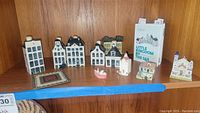 Shelf display of 7 Delft blue and white miniature Dutch houses, ceramic lighthouse, small pink boat, decorative tile, and book 'Little Kingdom by the Sea'.