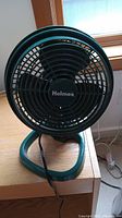 Front view of Holmes black desk fan with circular grille and base on wooden surface near window.