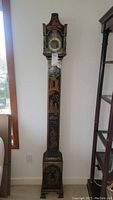 Full front view of the tall grandfather clock showing its slim black body with gold and hand-painted Asian-inspired figures and decorations.