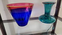 Two artisan hand blown glass vases on a glass shelf; one blue and pink with yellow rim, one teal with reddish rim and wider base.