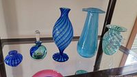 Six handblown glass items displayed on a glass shelf including blue and turquoise scent bottles and vases with varied shapes and patterns.
