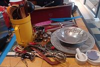 Full view of all kitchen and bar utensils including wooden spatulas in plastic container, pewter bowls, salt and pepper shakers, and various tools spread on table.