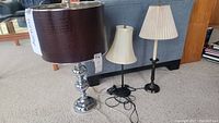 Photo showing three table lamps on carpeted floor including a large metallic base lamp with dark leather-like shade and two smaller lamps with pleated fabric shades.