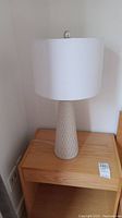 Single 27 inch beige textured ceramic table lamp with white round shade on wooden nightstand.