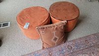 View of two round rust-orange ottomans and a square matching throw pillow sitting on carpet, showing fabric texture and color.