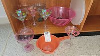 Photo showing six plastic stemware glasses (five colorful patterned, one pink), pink salad bowl with white lid, orange strainer.