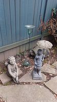 Photo showing a concrete cherub statue seated on the ground, a plastic girl statue holding an umbrella, and a glass and metal planter with a solar light spike.