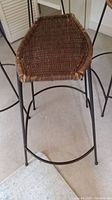 Close-up angled photo of a single stool showing dark brown rattan woven seat and black metal base with curved legs and footrest ring.