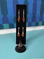Full display of two pairs of amber drop earrings and one amber ring on a black holder against a patterned background.
