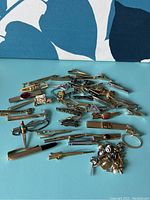 Wide arrangement of various men's tie clips and pins in metal finishes on blue background, showing design variety including tool shapes, knives, vehicles, initials and decorative stones.