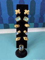 Jewelry display showing five pairs of gold-toned clip earrings with blue stones in various shapes mounted vertically on a black stand.