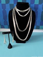 Front view of three layered sterling silver strand necklaces on black bust and drop earrings on stand against blue background.