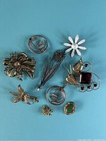 Lot of 9 sterling silver pins and brooches arranged on blue surface, showing variety of shapes and designs including floral and abstract geometric forms.