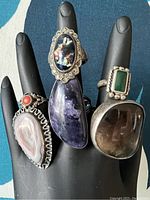 Rings displayed on a black mannequin hand showing front view of five rings with varied stones including agate, amethyst-like, green, smoky quartz, and iridescent stone.