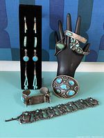 Photo showing multiple earrings mounted on black stand and three cuff bracelets plus a wide chain bracelet laid flat, all with turquoise stones and silver-tone metal.