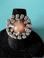 Front view of Rachel Gera sterling silver modernist ring with round pink stone on black display stand against turquoise background.
