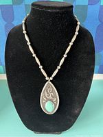 Front view of necklace and pendant displayed on black jewelry bust, showing pendant's engraved design and turquoise stone.