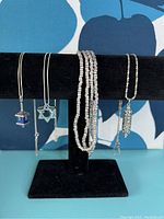 Four sterling silver Judaica-themed necklaces displayed on black stand, showing pendants including dreidel, Star of David, and Torah scroll with an additional multi-strand beaded necklace.