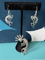 Photo shows two sterling silver Kokopelli earrings and a Kokopelli pendant on a black display stand against a blue and white background.