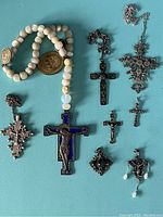Photo shows eight sterling silver crosses of different styles and sizes laid out on a blue surface along with a string of white rosary beads with attached medallions and one large blue enamel crucifix.