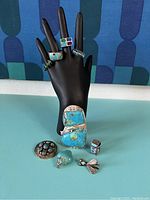 Photo showing multiple turquoise rings and other items displayed on black ring holder hand and turquoise surface