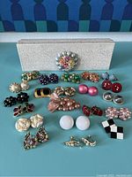 Photo showing 42 vintage clip earrings and brooches arranged on a green surface. Includes floral brooch, faux pearl earrings in various colors, geometric and bow-shaped clip earrings, and checkered patterned earrings.