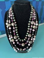 Four vintage cloisonné necklaces displayed on a black mannequin bust, showing various sizes and colors of beads with floral cloisonné decorations and solid beads in purple, pink, green, and black hues.