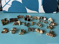 Photo showing multiple pairs of vintage men's cuff links arranged on a turquoise surface revealing a variety of gold tone and a few silver-tone and black enamel designs, confirming quantity and diverse styles
