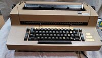Front view of beige-brown IBM daisy wheel typewriter showing keyboard, carriage, and branding plate.