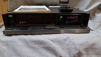 Front view of the Sony SLV-R5UC S-VHS VCR showing the power on with display active, front panel buttons and jog dial, along with remote and manual on top.