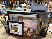 View of five framed art prints stacked together, showing different frame styles and partial image views including a scenic coastal sunset and muted traditional landscape artwork