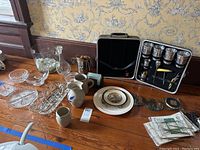 Photo showing an assortment of glassware, ceramic crockery pieces including jugs and mugs, a portable bar kit with shakers and bar tools, and small plates on wooden surface.