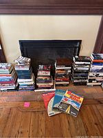 Wide view showing multiple stacks of books in front of a fireplace. Books of various sizes are stacked on the hearth and floor.