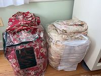 Two comforter sets placed side by side on a wooden floor; red floral set in packaging and cream floral set partly in a plastic cover.