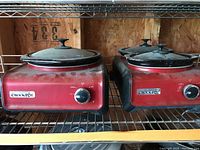 Two Crock-Pot red and black catering style food warmers with glass lids shown on metal shelving, dusty and used condition