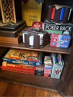 Photo showing various board games stacked on a wooden shelf including Advance to Boardwalk, Risk Storm Rising, Stratego, computer chess game, and card games, with a Book of 1000 Family Games and poker chips in revolving holders visible.