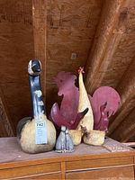 Photo shows large wooden goose figurine with two metal red rooster silhouettes, two smaller painted bird figurines, and two small cat figurines on a wooden surface under attic roof.