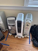 Three space heaters side-by-side on wooden floor; Lasko on left, two DeLonghi on right.