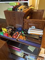 Large collection of office supplies on wooden shelves including staplers, hole punches, paper clips, glue sticks, scissors, pens, and envelopes.