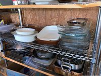 Wide shot showing metal shelving rack filled with various bakeware items including glass dishes, ceramic bakeware, metal roaster pans, springform pan, and cookware.