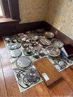 Wide view showing large assortment of silver-plated items laid on newspaper on wooden floor in corner of room with wallpaper.